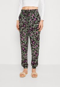 Los pantalones negros con flores presentan patrones en púrpura y verde, con una cintura elástica y puños fruncidos. Se complementan con sandalias trenzadas beiges.