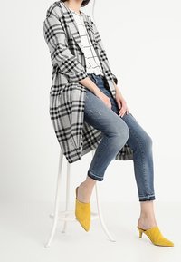 Mujer sentada en un taburete blanco con un abrigo de cuadros blanco y negro, una blusa a cuadros blanca, jeans azules y tacones amarillos.