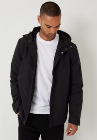 Sort vandtæt jakke med hætte, lynlås, trykknapper og sidelommer. Mat finish og justerbare manchetter. Bæres over en hvid t-shirt.