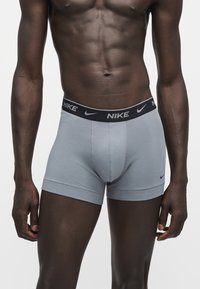 Μυώδης άνδρας που φορά ανοιχτό γκρι boxer της Nike με μαύρο λάστιχο στη μέση και λογότυπο, στέκεται σε απλό φόντο.