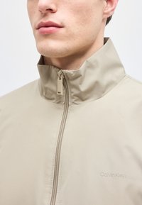 Μπεζ μπουφάν με φερμουάρ και ψηλό γιακά που φοράει ένα άτομο, με διακριτικό λογότυπο "Calvin Klein" στο στήθος.