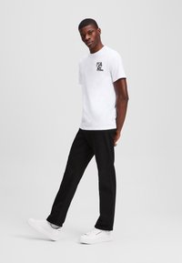 Witte T-shirt met zwart "KARL" logo, gecombineerd met zwarte broek en witte sneakers. Model staat in een neutrale achtergrond, kijkt recht vooruit.