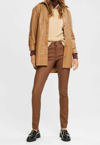 Hnědá koženka ve stylu košile s dvěma předními kapsami, pod ní béžový žebrovaný svetr a hnědé slim-fit kalhoty, doplněné černými lesklými botami.