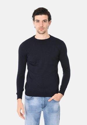 Uomo con capelli scuri che indossa un maglione nero aderente a maniche lunghe e jeans azzurri, in piedi con una mano in tasca.