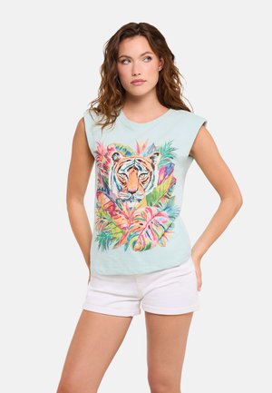 Mujer con camiseta sin mangas azul claro con estampado colorido de cara de tigre y hojas tropicales, combinada con pantalones cortos blancos, de pie con las manos en las caderas.