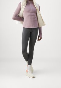 Person står iført en mauve langærmet top, en off-white ærmeløs jakke, mørkegrå leggings, hvide sokker og hvide sneakers med orange detaljer.