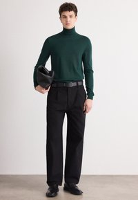 Grüner Rollkragenpullover kombiniert mit hoch geschnittenen schwarzen Hosen und einer schwarzen Tasche. Das Outfit zeichnet sich durch eine schlanke Passform und einen strukturierten Stoff aus.