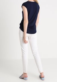 Kvinna iklädd en marinblå kortärmad topp, vita slim-fit byxor och ljusblå klacksandaler, stående framför en helt vit bakgrund.