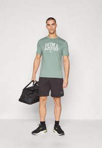 Grünes T-Shirt mit kurzen Ärmeln und weißem "PUMA Sports Club"-Druck, kombiniert mit schwarzen Shorts und schwarzen Sneakers, während eine schwarze Sporttasche gehalten wird.