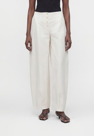Femme portant un pantalon blanc taille haute à jambes larges avec des rayures verticales noires et des boutons, associé à un haut noir sans manches et des sandales noires à talons.