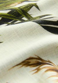 Textil mit einem hellen Creme-Hintergrund und grünen sowie braunen Blättermustern. Der Stoff hat eine weiche, strukturierte Oberfläche mit sichtbaren Fasern.