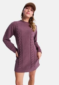 Nainen, jolla on päällään violetti palmikkoneulemekko ja vaaleanbeige hattu, poseeraa kädet taskuissa valkoista taustaa vasten.