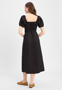 Schwarzes Midi-Kleid mit quadratischem Rückenausschnitt, gerafften Puffärmeln und einem gerafften Rückenteil, hergestellt aus einem strukturierten Stoff. Braune Slipper.