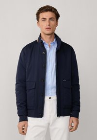 Chaqueta azul marino con cuello alto, cierre de botones, dos bolsillos frontales y puños elásticos, llevada sobre una camisa de color azul claro.