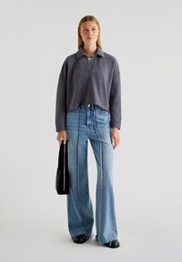 Sweatshirt i mørkegrå med krave, parret med lyseblå flared jeans med en flosset kant og sorte sko. Holder en sort tote bag.