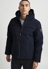 Chaqueta con capucha de color marino con diseño acolchado, cremallera frontal, dos bolsillos decorativos y un parche con el logotipo en la manga izquierda. Tejido ligero.