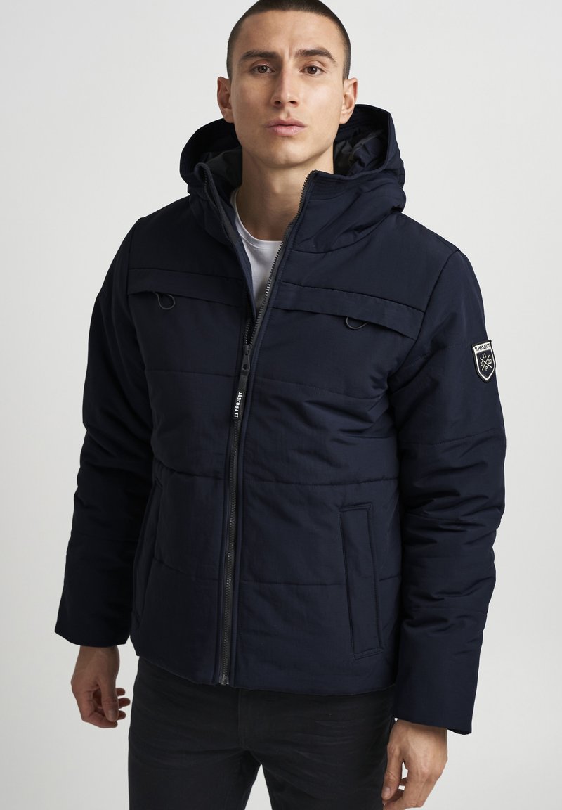 Chaqueta con capucha de color marino con diseño acolchado, cremallera frontal, dos bolsillos decorativos y un parche con el logotipo en la manga izquierda. Tejido ligero.