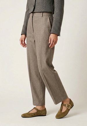 Person trägt braune, hochgeschnittene, maßgeschneiderte Hose, graue Fischgrätenjacke und olivgrüne Veloursleder-T-Riemchen-Schuhe mit Schnalle.