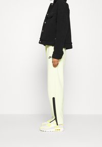 Black fleece jacket with button front, worn over light yellow sweatpants featuring a black Nike logo and side zippers, paired with white and yellow sneakers.