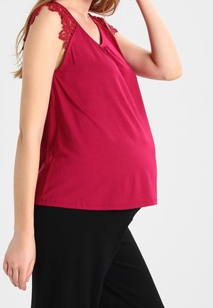 Gravid kvinde iført en ærmeløs rød top med blonder på skuldrene og sorte bukser, poserer med den ene arm hævet.