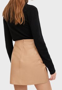 Mini-jupe en cuir fauve avec une texture lisse et des lignes épurées, dotée d'une fermeture éclair au centre du dos et de coutures minimales. Haut noir en partie visible.