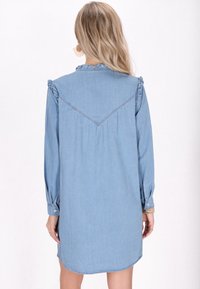 Svetlomodrá džínová košeľová šaty s dlhými rukávmi, volánovými detailami na pleciach a zakriveným lemom. Má golier a manžety na gombíky.