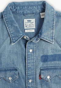 Jasnoniebieska koszula dżinsowa o luźnym kroju, z ostrym kołnierzykiem, zatrzaskami, dwiema kieszeniami na piersi i metką Levi's. Widoczne szczegóły ściegów.