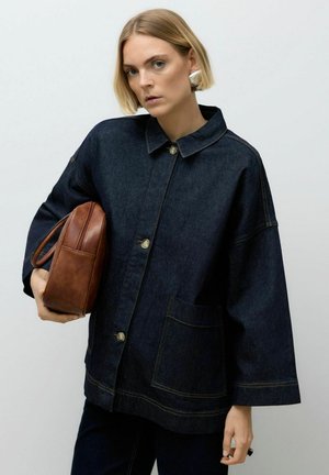 Giacca di denim scuro con colletto, bottoni frontali e una grande tasca. Il modello tiene una borsa di pelle marrone, lo sfondo dell'impostazione è neutro.