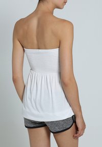 Weißes trägerloses Oberteil mit gerafftem Mieder und ausgestelltem Saum, kombiniert mit grauen Shorts mit schwarzem Besatz. Glatte Textur und leichtes Material.