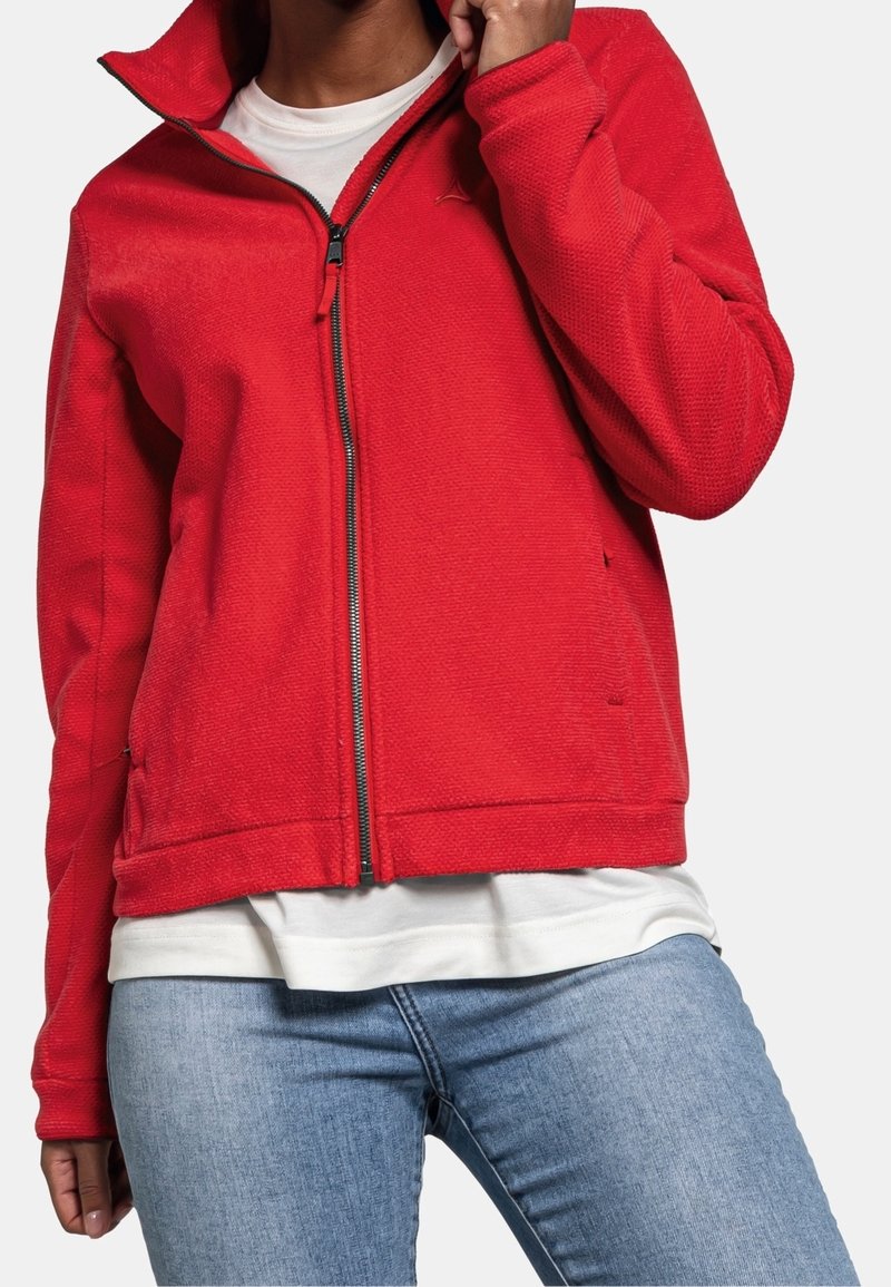 Personne portant une veste rouge zippée sur une chemise blanche et un jean bleu clair, avec la main près du menton et la veste partiellement dézippée.