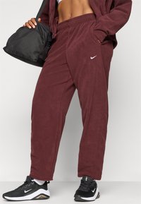 Bordové flísové jogger nohavice so bočnými vreckami a elastickým pásom, s malým bielym logom Nike; v kombinácii s čiernymi športovými topánkami.