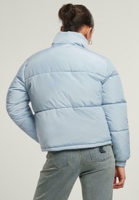 Lysblå dunjakke med højt krave, der har quiltede sektioner og en glat, blank overflade. Bæres med højtaljede denimjeans.