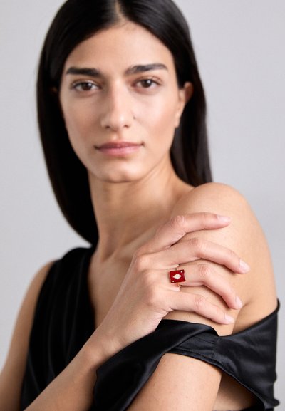 Červený diamantový prsteň s obdĺžnikovým dizajnom, zlatým pásikom a malými ozdobami. Ruka modelky spočíva na holých ramenách, oblečená v čiernom topu.
