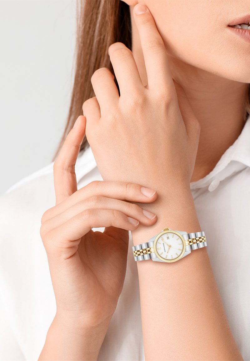 Montre ronde avec un cadran blanc, des accents dorés et un bracelet en métal argent et or. Portée sur une main à la peau claire.