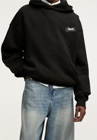 Czarna bluza z kapturem z przednią kieszenią i wyszywanym białym logo. Połączona z jasnoniebieskimi jeansami o luźnym kroju i wyblakłej fakturze.