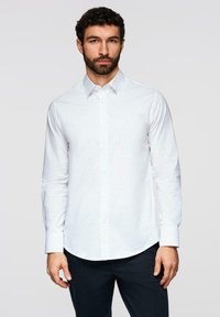 Homme aux cheveux foncés et à la barbe portant une chemise blanche à manches longues à petits pois et un pantalon sombre, debout devant un fond gris.