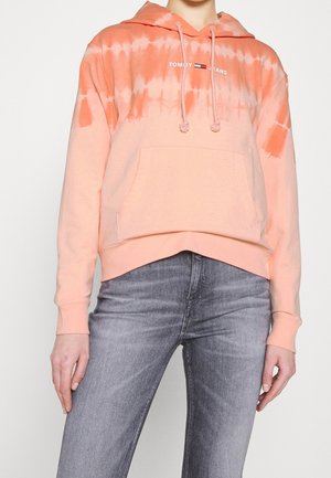 Sudadera con efecto tie-dye en melocotón, con un bolsillo frontal y cordones; presenta el logo de la marca en blanco. Combinada con jeans de mezclilla gris claro.