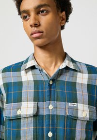 Camisa de cuadros en colores azul, verde y blanco. Cuenta con dos bolsillos frontales, botones y un cuello. Hecha de tela texturizada. Logotipo de Wrangler presente.