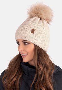 Căciulă tricotată crem cu un model texturat, având un pompon mare din blană sintetică în vârf și o mica etichetă din piele pe mansetă.