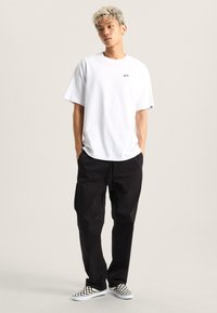 White cotton t-shirt with a small logo, paired with black drawstring pants and black-and-white checkered slip-on shoes.