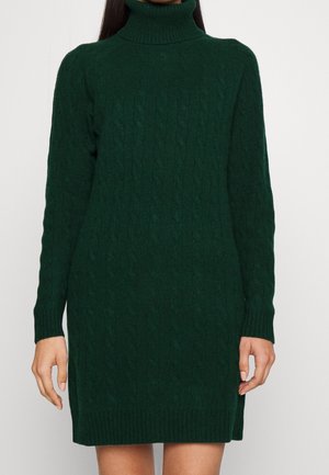 Žena v tmavě zelených svetrových šatech s rolákem a dlouhými rukávy stojí před jednoduchým světlým pozadím.