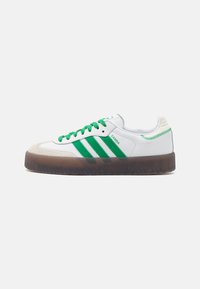 Sneaker blanc avec des accents verts, présentant trois bandes, des lacets verts et une semelle en caoutchouc brun. Capuchon de nez en daim distinctif et talon texturé.