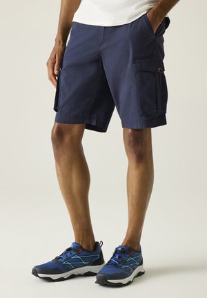 Man die marineblauwe cargoshorts en blauwe sportschoenen draagt, staand met één hand in de zak tegen een neutrale achtergrond.