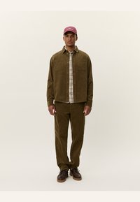 Olivgrüne Cordjacke mit zwei Brusttaschen, über einem karierten Hemd getragen. Passende Cordhosen und braune Schnürschuhe.