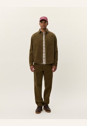 Olive green corduroy shirt jacket with two chest pockets, layered over a plaid shirt. Matching corduroy pants and brown lace-up shoes.