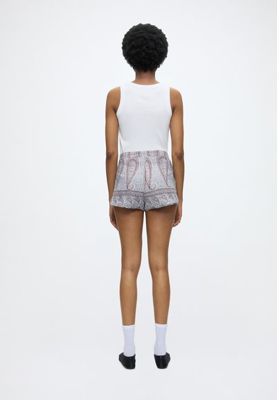 Femme aux cheveux courts et bouclés portant un haut blanc sans manches, un short à motifs avec ourlet volanté, des chaussettes blanches et des chaussures noires à enfiler, debout, de dos.