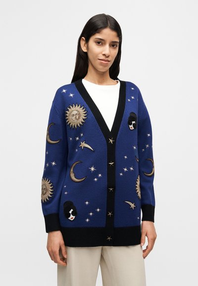 Cardigan en tricot bleu avec des accents noirs, présentant des motifs célestes brodés, incluant des soleils, des lunes, des étoiles et un détail de visage.