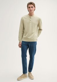 Jasno beżowa koszula z długim rękawem i dekoltem henley oraz trzema guzikami, zestawiona z niebieskimi jeansami i jasnobrązowymi trampkami. Gładka tekstura.