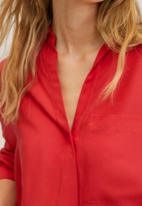 Donna con lunghi capelli biondi che indossa una camicia rossa con bottoni e taschino, mostrata dalla gola fino a metà torso su uno sfondo chiaro.