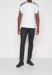 Camiseta atlética blanca con rayas negras en los hombros, combinada con pantalones negros que tienen rayas blancas a los lados y un logo, usados con zapatos negros.